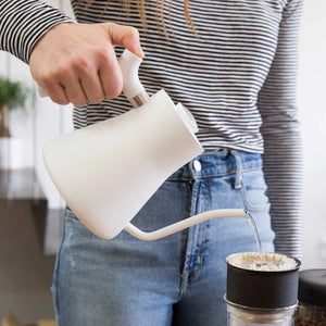 Roka, kas lej ūdeni no baltas ūdenskannas ar šauru snīpi uz kafijas filtru, daļa no EspressoGear, viss par kafiju.